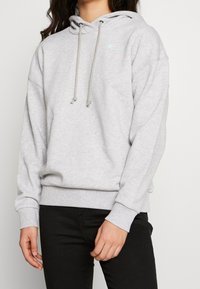 Šedá mikina s kapucí a šňůrkami, dlouhými rukávy, žebrovanými manžetami a lemem. Má malou barevnou nášivku na hrudi. Měkká textilie.