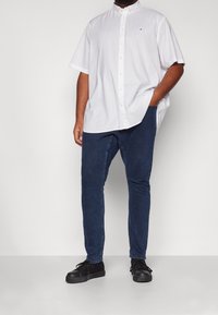 Uomo che indossa una camicia bianca a maniche corte con bottoni, jeans slim-fit blu scuro e sneakers nere, in piedi con una mano in tasca.