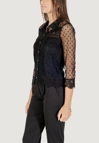 Blusa nera con bodice in pizzo floreale e maniche trasparenti a pois. La camicia ha una chiusura con bottoni e un orlo in pizzo ondulato.