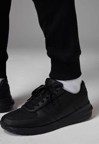 Black athletic shoes with textured leather and mesh upper, featuring a flat sole and traditional lacing. Worn with white ribbed socks.
