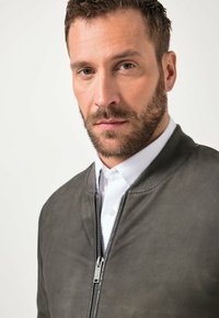 Baardige man met kort bruin haar, gekleed in een grijze jas met rits over een wit overhemd met kraag, tegen een effen lichte achtergrond.