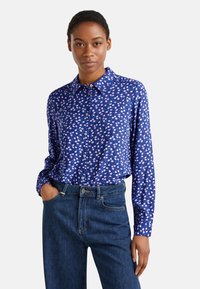 Camicia a maniche lunghe blu con motivo floreale in rosa, bianco e verde. Presenta un colletto button-down ed è abbinata a jeans in denim a vita alta.