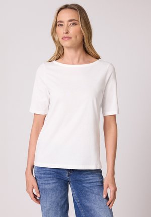 Mujer con cabello largo y rubio, vestida con una camiseta blanca lisa de manga corta y jeans azules, de pie frente a un fondo claro.