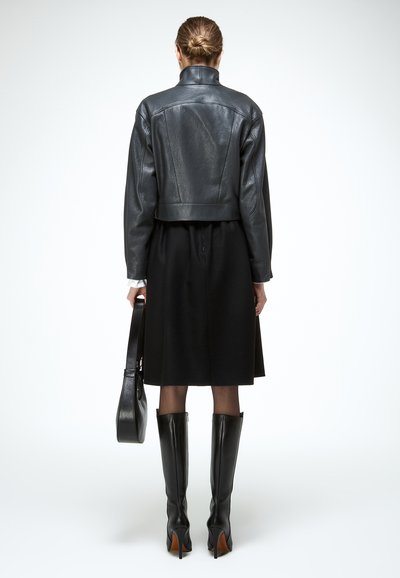 Vrouw met opgestoken haar, zwarte leren jas, zwarte rok, kniehoge zwarte laarzen en een zwarte handtas in haar hand, met haar rug naar voren op een witte achtergrond.