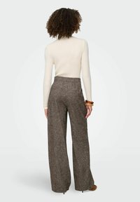 Kobieta z kręconymi włosami, ubrana w kremowy prążkowany sweter z długim rękawem, brązowe szerokie spodnie o fakturze i brązowe szpilki, widziana z tyłu.