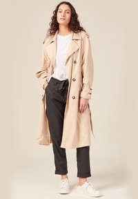 Beige trenchcoat med stora knappar, bred krage och knytmidja. Kombinerad med mörkgrå byxor och vita sneakers, mot en neutral bakgrund.