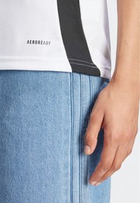 Bijela i crna sportska majica s teksturiranim materijalom, koja sadrži "AEROREADY" logotip. Svijetlo plave traperice kratke hlače s istaknutim vertikalnim šavovima.