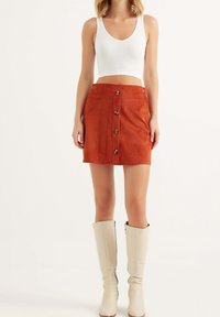 Femme portant un top court blanc, une jupe en velours côtelé couleur rouille avec boutons sur le devant, et des bottes beige montantes jusqu'aux genoux, debout devant un fond uni.