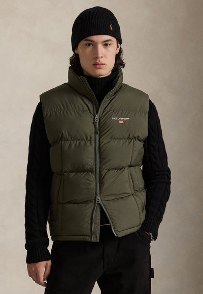 Gilet imbottito verde oliva con cerniera frontale, collo alto e logo "POLO SPORT"; indossato sopra un maglione nero a trecce.