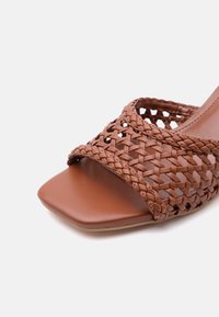 Brązowy skórzany sandał z kwadratowym, otwartym noskiem i plecionym paskiem na przodzie na białym tle.