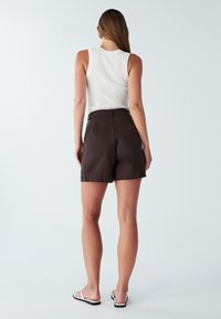 Bruine hooggewaisted shorts met een gladde textuur en zijzakken, gecombineerd met een witte geribbelde tanktop. Sandalen hebben zwarte zolen.