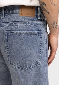 Ξεθωριασμένο μπλε τζιν με καφέ ετικέτα στη πίσω μέση. Διαθέτει κλασικό σχέδιο με πέντε τσέπες και εμφανείς ραφές.