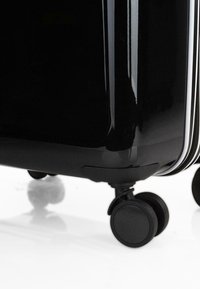 Maleta rígida negra con un acabado brillante, equipada con un mango retráctil, ruedas negras suaves y acentos en cremallera blanca.