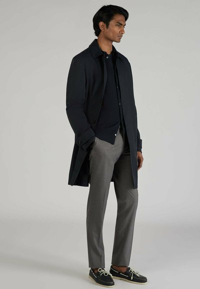 Cappotto lungo nero sopra un maglione blu navy, abbinato a pantaloni grigi e scarpe scure. Presenta polsini abbottonati e una vestibilità su misura.