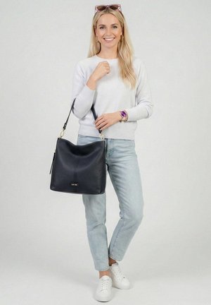 Naeratav naine, kellel on pikad blondid juuksed, kannab heledat kampsunit, teksapükse, valgeid tossusid, lillat kella, päikeseprille ning hoiab suurt mustast nahast käekotti.