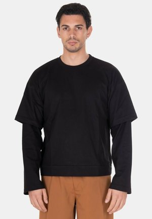 Giovane uomo con capelli corti e scuri indossa una camicia nera a strati con maniche lunghe e corte e pantaloni marroni, in piedi davanti a uno sfondo bianco.