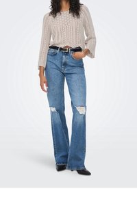 Beige gehaakte top met lange klokmouwen, gecombineerd met lichtblauwe gescheurde wijde jeans en zwarte puntige schoenen. Zwarte riemdetails.