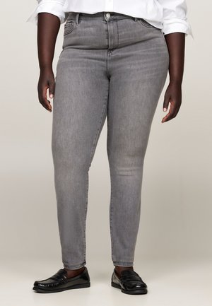 Person iført grå slim-fit jeans, sorte loafers og en hvid skjorte med opkrogede ærmer, stående mod en ensfarvet baggrund.