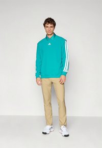 Homem jovem vestido com sweat verde-azulado com riscas brancas nas mangas, calças bege e ténis brancos, em fundo cinzento liso.