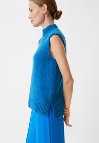 Blaues ärmelloses Strickoberteil mit hohem Kragen, das mit einer strukturierten Oberfläche und einem gerippten Saum versehen ist, kombiniert mit einem fließenden blauen Rock.