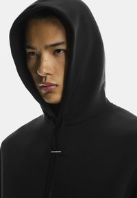 Zwarte hoodie van zacht materiaal, met een grote capuchon, trekkoorden en een klein merklabel aan de voorkant. Close-up van de halslijn en het gezicht.