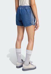 Osoba ubrana w niebieskie dżinsowe szorty z białymi paskami po bokach, szare skarpety Adidas do kostki oraz szare sneakersy Adidas, stojąca na jasnym tle.