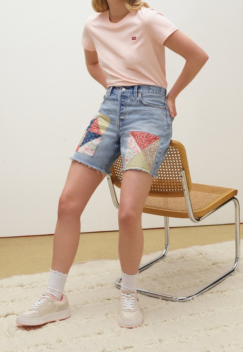 Personne portant un t-shirt rose clair et un short en jean taille haute avec un patchwork coloré, debout près d'une chaise tissée sur un tapis texturé.