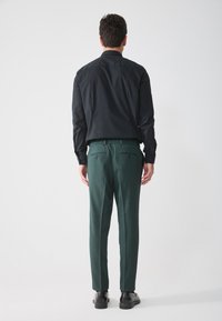 Schwarzes Langarmshirt und dunkelgrüne Hose mit elastischem Bund. Die Hose verfügt über zwei Gesäßtaschen. Modell von hinten gezeigt.