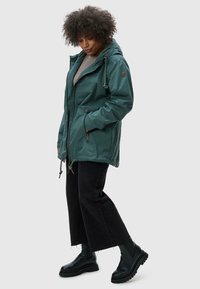 Groene waterdichte jas met capuchon, trekkoord in de taille en zijzakken. Draagt over een grijze top, in combinatie met zwarte cropped broek en laarzen.