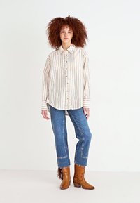 Camicia a righe beige e bianca, abbinata a jeans in denim blu. Il modello indossa stivali da cowboy beige con dettagli in frappa.