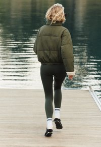 Pikowana oliwkowa kurtka z kołnierzem z polaru, czarne legginsy, białe skarpetki w paski i czarne buty. Osoba odchodzi na drewnianym pomoście.