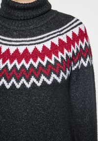 Dzianinowy sweter z golfem w ciemnoszarym kolorze z wzorzystą wstawką, która ma zygzakowate wzory w czerwonym, białym i czarnym kolorze. Miękka tekstura.