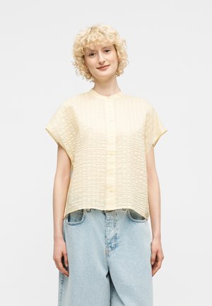 Nainen lyhyillä kiharoilla vaaleanruskeilla hiuksilla, joka pukeutunut vaaleankeltaiseen, lyhythihaiseen kuvioituun paitaan ja vaaleansinisiin korkeavyötäröisiin farkkuihin.