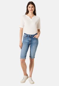 Maglietta corta bianca con abbottonatura; bermuda in denim blu con risvolti; sneakers beige con lacci.