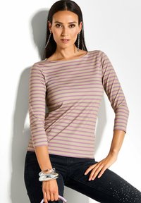 Vrouw in een roze en beige gestreepte driekwarts mouwtop, zwarte broek met versieringen, en een lavendelkleurige tas met zilveren armbanden.