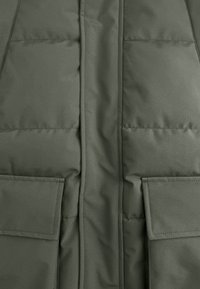 Close-up van de groene gewatteerde jas voorkant met de centrale naad en rechthoekige klepzakken met zichtbare stiksels.