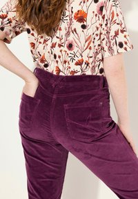 Pantalons en velours côtelé violets avec une surface texturée, dotés de poches arrière et d'une taille haute, associés à un haut à motifs floraux.