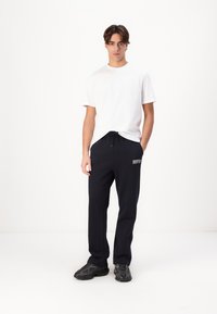 Weißes T-Shirt mit kurzen Ärmeln, kombiniert mit navyblauen Jogginghosen mit Kordelzug und gesticktem Logo, ergänzt durch dunkle Sneaker.