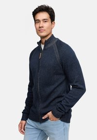 Granatowy rozpinany sweter z wysokim kołnierzem, ściągaczami na rękawach i teksturowanym wzorem dzianiny. Posiada drewniany uchwyt suwaka jako detal.