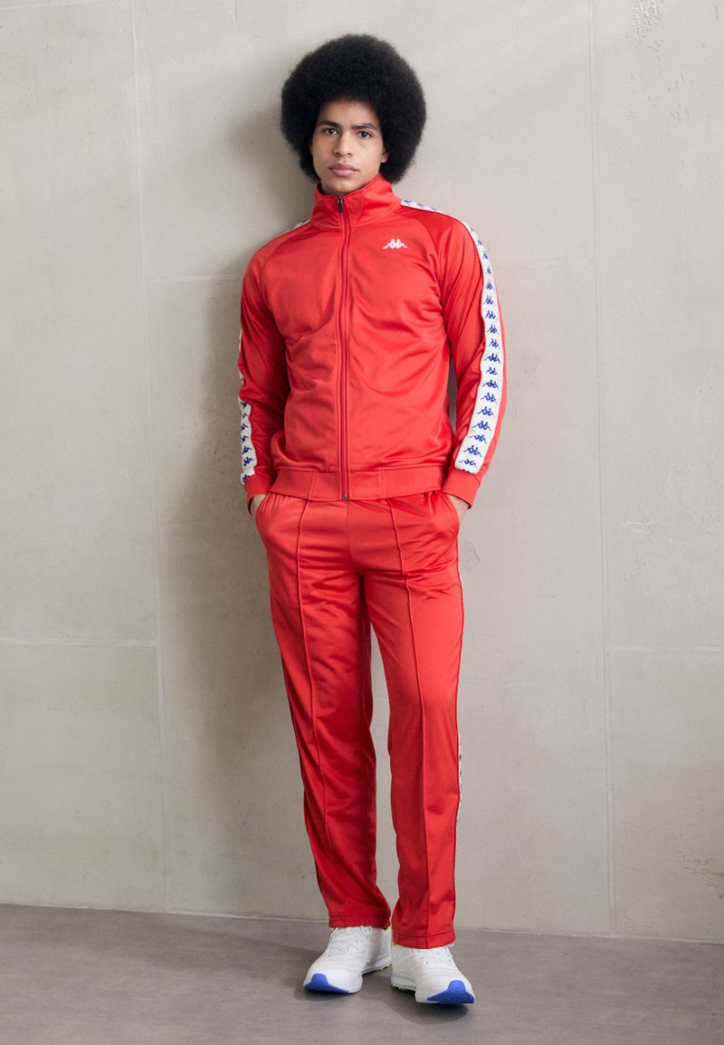 Survêtement rouge en polyester, comprenant une veste zippée avec des accents en motifs blancs et bleus sur les manches, assorti à un pantalon slim.
