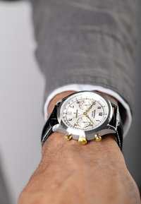 Silver chronograph watch with a white dial, black leather strap, gold accents, and three sub-dials. Worn on a wrist with a textured background.
