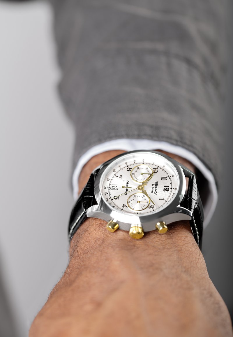 Silver chronograph watch with a white dial, black leather strap, gold accents, and three sub-dials. Worn on a wrist with a textured background.