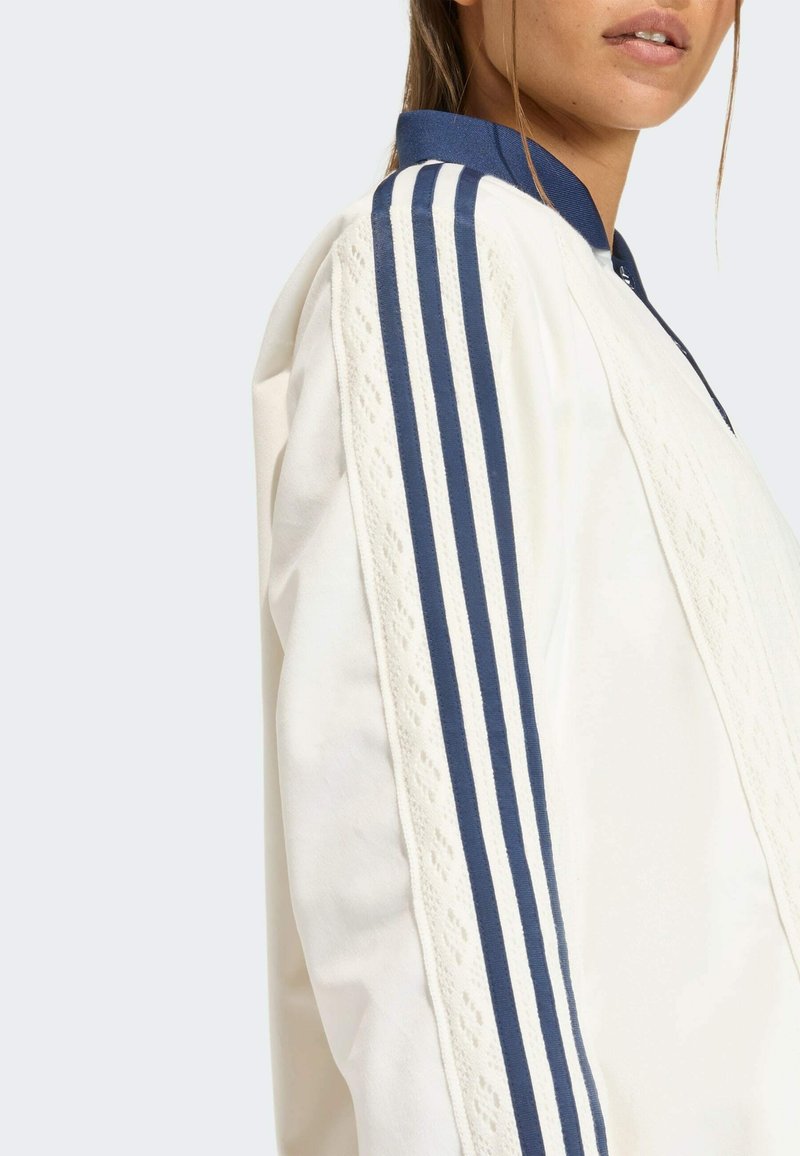 Hvid atletikjakke med marineblå accenter og tre parallelle striber på ærmerne. Har et tekstureret mønster langs skuldrene.