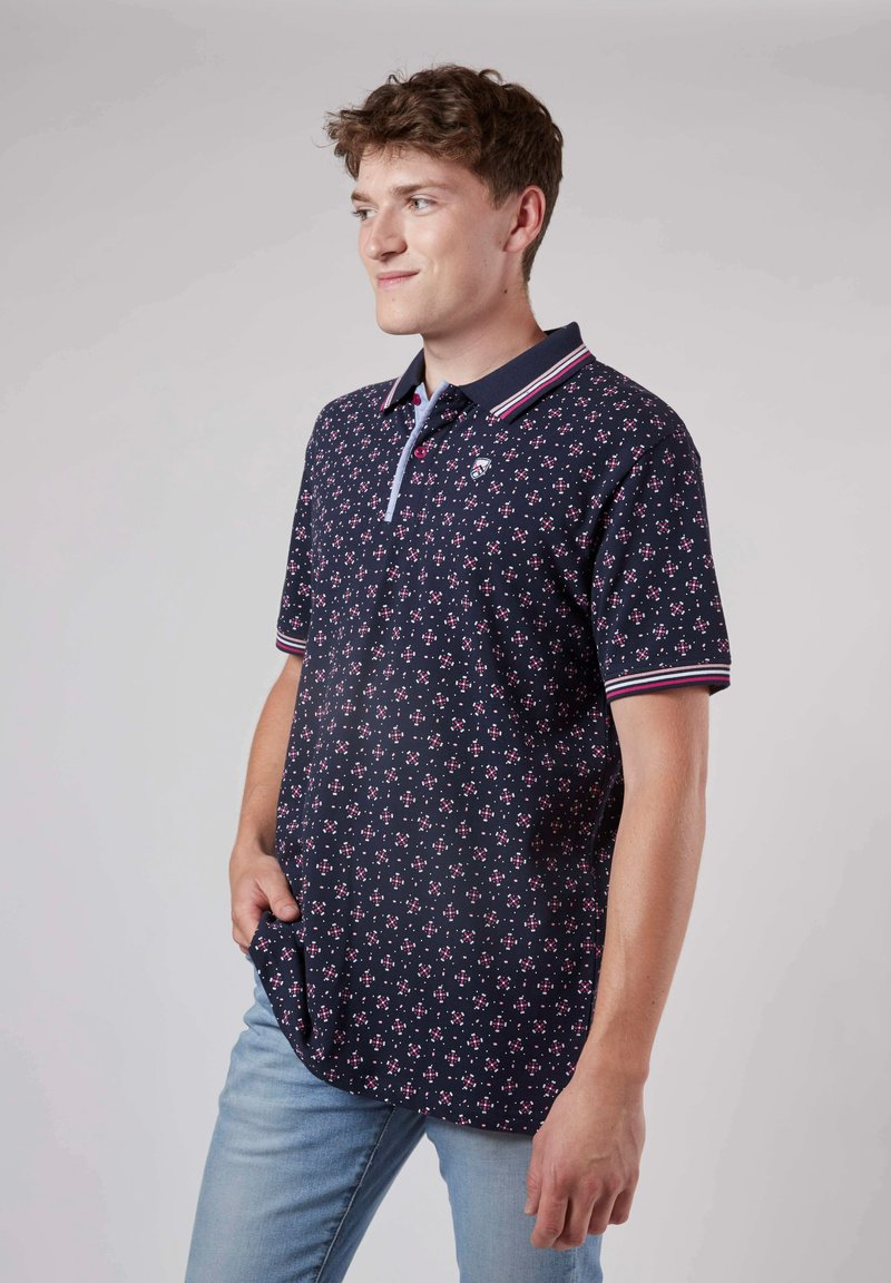 Polo shirt marine avec un motif floral rose et blanc, doté d'un col à boutons et d'accents rayés sur les manches. Tissu en mélange de coton doux.