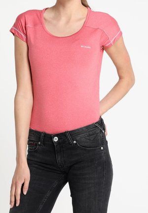 Vrouw draagt een aansluitend roze top met korte mouwen en zwarte high-waisted jeans, staat met één hand op de heup en de andere langs haar zijde.