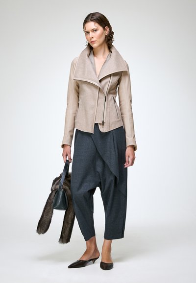 Femme portant une veste zippée asymétrique beige et un pantalon ample gris foncé, tenant un sac à main noir avec un accessoire en fourrure sur fond uni.