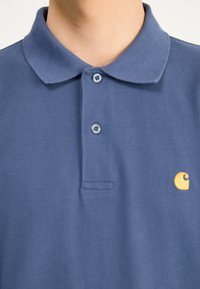 Tumesinine puuvillane polo särk, millel on krae, kaks nuppu ja vasakul rinnal kollane logo. Tekstureeritud kangas peene mustriga.