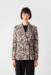 Vrouw in een beige dubbelzijdig jasje met zwart en bruin luipaardprint over een zwarte top en zwarte broek, staand tegen een effen achtergrond.