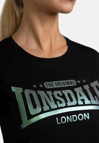 Juoda marškinėliai su "LONSDALE" metalinės sidabro spalvos spauda, apvaliu kaklu ir trumpomis rankovėmis, matomi nuo pečių iki viršaus.