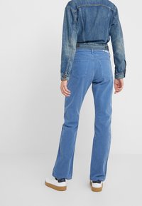 Personne portant un pantalon en velours côtelé bleu, une veste en jean et des baskets blanches, debout de dos sur un fond blanc uni.